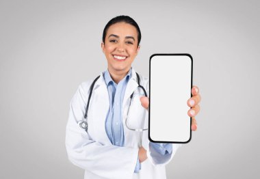 Gülümseyen Brezilyalı kadın doktor kameraya boş ekranlı akıllı telefon gösteriyor, gri arka planda tek başına duruyor, maket. Latin terapist kadın çevrimiçi tıbbi teklif öneriyor