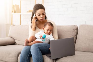 Siyah anne bebeğe birden fazla iş veriyor, telefonla konuşuyor ve evdeki koltukta dizüstü bilgisayar kullanıyor. Afrikalı Amerikalı serbest çalışan anne doğum izni sırasında uzaktan çalışıyor. Annelik Konsepti