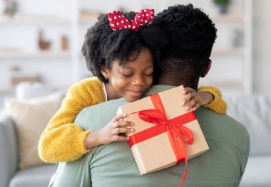 Gülümseyen küçük siyah kız, hediye kutusunu tutuyor, babasını kucaklıyor, babasını babasıyla selamlıyor, doğum gününde ya da doğum gününde, mutlu Afro-Amerikan ailesi evde kaynaşıyor, yakın çekim.