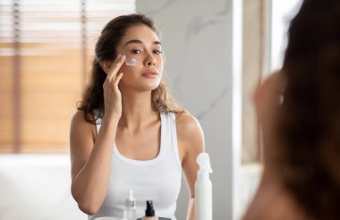 Woman Applying Facial Moisturizer Cream On Face Touching Cheeks In Bathroom At Home. Female Skincare Routine And Cosmetics, Beauty And Pampering Concept. Selective Focus
