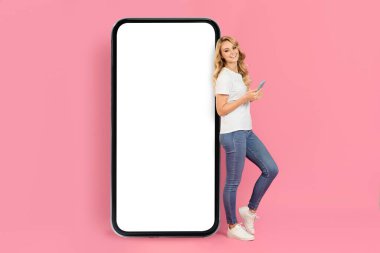 Uzun sarı saçlı genç bir kadın akıllı telefonu tutarken gülümsüyor. Parlak pembe bir arka planda büyük, boş bir akıllı telefona yaslanıyor..
