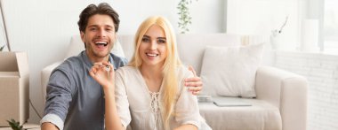 Genç bir çift, parlak oturma odalarındaki rahat bir kanepede otururken sıcak bir şekilde gülümsüyor. Etrafı bitkilerle ve kutularla çevrili. Mutluluk ve bağlantı anlarını sergiliyorlar.