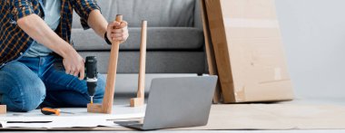 Mobilya evi. Milenyum çocuğu mobilya tamir ediyor, ahşap masayı tamir ediyor elektrikli matkap kullanıyor, evde oturuyor, tornavidayla vidalanıyor ve oturma odasındaki dizüstü bilgisayara bakıyor.