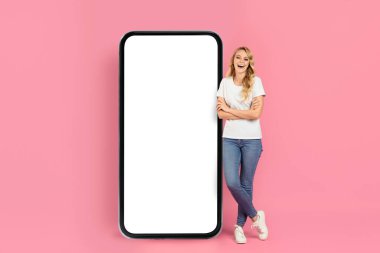 Genç bir kadın büyük boş bir akıllı telefon ekranının yanında duruyor, parlak pembe bir stüdyoda güvenle gülümsüyor. Günlük kot pantolon ve tişört giyiyor, neşeli bir atmosfer yaratıyor..
