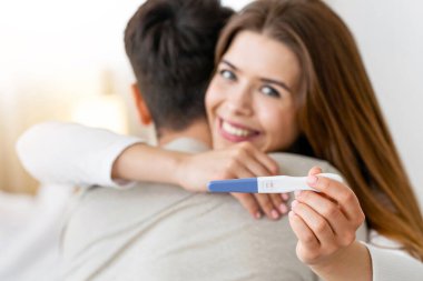 Focus on pregnancy test. Young husband and wife hugging at home clipart