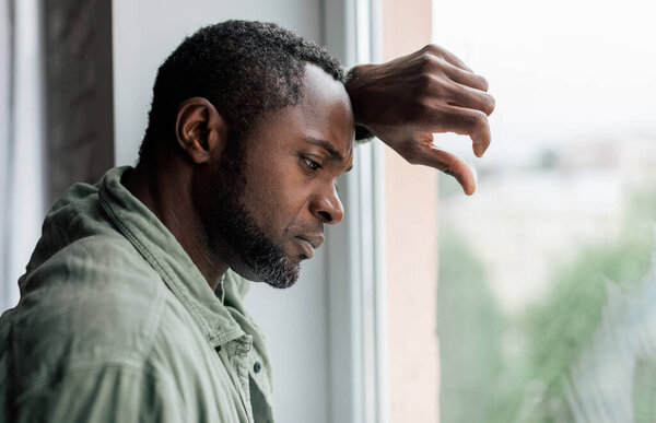 Unhappy frustrated adult african american guy in casual suffering from depression and bad news near window in home interior. Health problems, stress from self-isolation during covid-19 quarantine