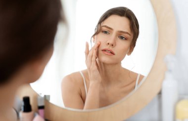 Sivilceye yatkın deri konsepti. Evdeki aynaya bakıp, cildini kontrol eden üzgün genç bir kadın.