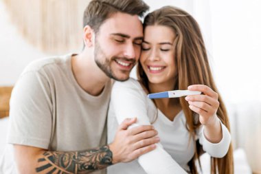 Haberler iyi. Neşeli adam kıza sarılıyor ve yatak odasında pozitif hamilelik testi gösteriyor.