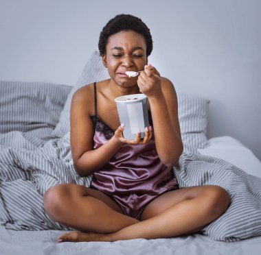 Breakup and sadness concept. Crying african american girl eating ice cream in bed at home clipart