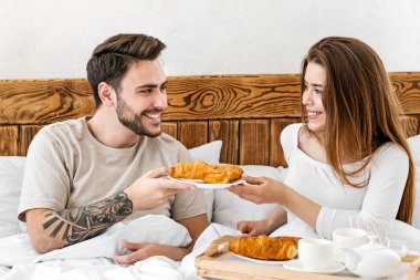 İki konsept için romantik kahvaltı. Dizlerinin üstünde tepsisi olan kız, yatakta adama kruvasan veriyor.