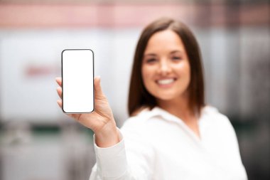 Bin yıllık bir iş kadını bir ofiste akıllı telefonu tutarken kendine güvenen bir şekilde gülümsüyor. Modern çalışma sahasında parlak mobilyalar ve işbirliği için ideal profesyonel bir atmosfer yer alıyor..