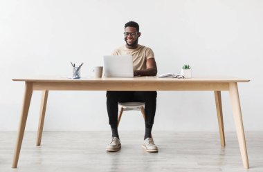 Happy young black man using laptop computer for online work at table in home office, free space. Cool African American guy having remote job, freelancing on web. Modern business concept