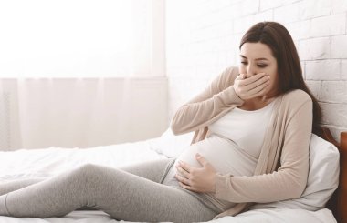 Sabah bulantısı. Ağzını hamilelik sırasında boş alan mide bulandırıcı hissi kapsayan yatağın üstüne oturan genç hamile kadın