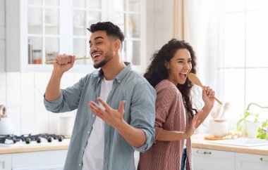 Ev Konseri. Neşeli Orta Doğulu eşler mutfakta eğleniyor, şarkı söylüyor ve dans ediyor. Neşeli Arap Çifti Spatulaları Mikrofon Olarak, Modern Müslüman Aile Evde Yemek Pişirirken Kandırıyor
