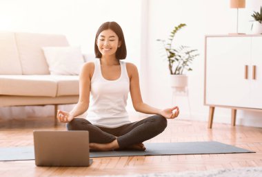 Sakin ol. Mutlu Asyalı kadın antrenörle internetten bilgisayar bağlantısı üzerinden meditasyon yapıyor. Nilüfer pozisyonunda yoga minderinde oturmuş bilgisayara bakıyor ve gülümsüyor.