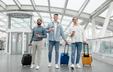 Vacation, Finally. Three Friends Men Traveling Abroad Walking With Suitcases And Holding Paper Coffee Cups Posing In Modern Airport Terminal Indoor. Tourism Concept. Full Length
