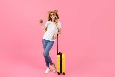 Genç bir kadın, elinde hindistan cevizi içeceği tutan parlak sarı bir valizin yanında güvenle duruyor. Güneş gözlüğü ve hasır şapka takıyor. Pastel pembe bir ortamda eğlenceli bir yaz kaçamağına hazır..
