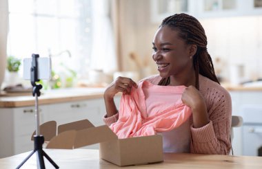 Boks videosu açılıyor. Afrikalı Kadın Moda blogcusu evdeki stil blogu için kayıt içeriği, üç ayaklı akıllı telefon, mutfakta oturan siyah kadın kamerada yeni kıyafetlerle birlikte paketleri açıyor.