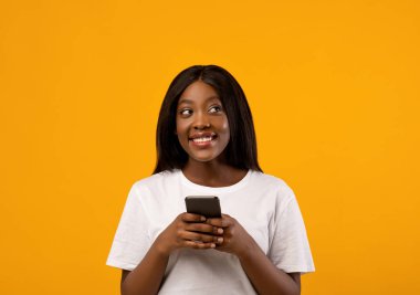 Gülümseyen Afro-Amerikan genç bayan cep telefonu kullanıyor ve yana bakıyor, kopya alanı, sarı stüdyo arka planı. Güzel siyah bayan akıllı telefon tutuyor, yeni ve güzel bir mobil uygulama kullanıyor.