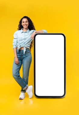 Büyük akıllı telefonun yanında boş ekranlı neşeli genç bir bayan, sarı stüdyo arka planında web sitesi, reklam ya da uygulamayı gösteriyor.