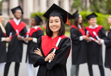 Mutlu Asyalı kız öğrenci mezuniyet partisi veriyor, mezuniyet kostümü giyiyor ve elinde diploma var, güzel bayan mezun üniversitede gururla poz veriyor, geçmişi bulanık.