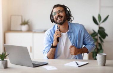 Keyifli Hintli bağımsız adam ev bilgisayarında çalışırken kulaklıkla müzik söylüyor ve dinliyor, akıllı telefonu mikrofon olarak kullanıyor, en sevdiği şarkının tadını çıkarıyor, uzay boşluğunu kopyalıyor.