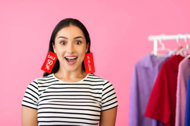 A young Caucasian woman displays excitement while shopping, wearing large sale tags in her earrings. She is surrounded by colorful clothes in a vibrant store, emphasizing consumerism. clipart