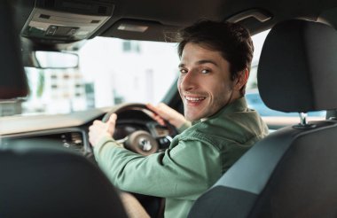 Yakışıklı genç adam kameraya bakıyor, arabada oturuyor, otomobili park ediyor, arka koltuğu görüyor. Modern bayilikte otomobil alan mutlu milenyum adamı, kopyalama alanı