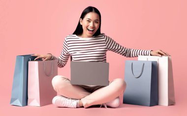 Çevrimiçi Alışveriş Konsepti. Dizüstü bilgisayarı ve parlak alışveriş çantaları olan neşeli Asyalı kadın pembe stüdyonun arka planında oturuyor, Koreli kadın satışlardan ve indirimlerden zevk alıyor, bedava yer var.