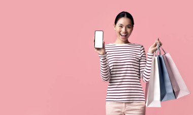 Heyecanlı Asyalı kadın beyaz ekranlı, elinde alışveriş torbalarıyla akıllı telefon gösterisi yapıyor. Neşeli Koreli alışveriş meraklısı kadın internetten alışveriş yapıyor. Pembe stüdyo arka planının başında duruyor.