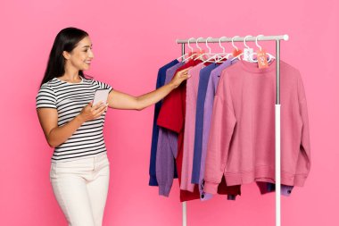 Neşeli bir suratı olan genç beyaz bir kadın canlı bir mağazada renkli tişörtleri karıştırıyor. Telefonundaki fiyatları kontrol ediyor, alışveriş cümbüşünün tadını çıkarıyor..