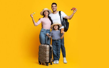 Küçük kızı tatile hazır mutlu bir Arap ailenin portresi, neşeli bir Orta Doğulu ebeveyn ve çocuklarının bavulları, pasaportları ve sarı arka planda seyahat hatıraları.