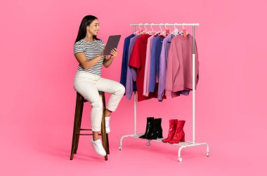 Genç beyaz bir kadın tabletinin üzerinde otururken etrafını renkli kıyafetlerle sarmış tabletini karıştırıyor. Parlak pembe arka plan neşeli alışveriş atmosferini güçlendirir.