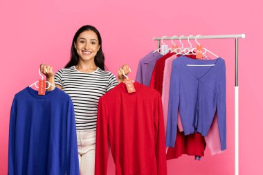 Genç bir kadın elinde parlak renklerde iki kazak tutarken gülümsüyor. Ek süveterlerle dolu bir rafın önünde duruyor, pembe bir arka planla çevrili..