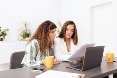 Parlak bir şirket ofisinde, iki kadın masa başında belgelerini tartışırken işlerine odaklanırlar. Kahve içerken fikirlerini paylaşıyorlar, takım çalışmasını ve üretkenliği vurguluyorlar..