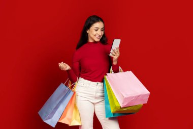 Neşeli bir yüzü olan genç bir kadın bir elinde renkli alışveriş çantaları, diğer elinde akıllı bir telefon tutuyor. Koyu kırmızı bir arkaplanda soyutlanmış, tüketime olan sevgisini gösteriyor..