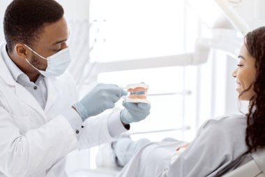 Diş hijyeni. Siyah diş hekimi çene mankeni tutuyor ve kadın hastaya diş fırçalamayı öğretiyor, profesyonel stomatolog modern klinikte genç Afrikalı Amerikalı kadınlara danışmanlık yapıyor,