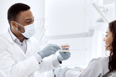 Genç siyahi stomatolog plastik çene tutuyor ve kadın hastaya diş hijyeni öğretiyor, dişçi genç kadına diş fırçalamayı öğretiyor Modern Klinikte kontrol sırasında dişlerini nasıl fırçalayacağını,