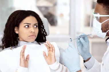 Klinikte kontrol sırasında şok geçiren diş hekimine bakan endişeli siyah kadın hasta diş tedavisi fobisinden muzdarip olan Afrikalı Amerikalı kadın elleriyle hareket etmeyi kesiyor.