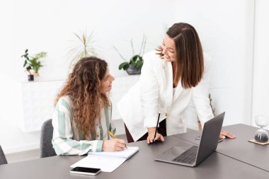 Çağdaş bir ofiste, iki kadın bir projede birlikte çalışır. Biri oturuyor, not alıyor, diğeri ise fikirleri tartışmak için öne eğiliyor. Bitkiler çalışma alanına taze bir dokunuş ekler..