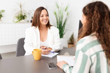 İki kadın modern bir ofis ortamında verimli bir konuşma yapıyorlar. Ceket giymiş bir kadın geri bildirim yaparken, diğeri not alıyor ve işbirliğini teşvik ediyor gibi görünüyor..