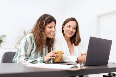 Parlak bir ofis ortamında iki kadın dizüstü bilgisayar üzerinde çalışırken fikirlerini tartışmaktan mutlu oluyorlar. Bir kahve fincanı, rahat ama odaklanmış bir takım çalışması atmosferi sergiliyor..