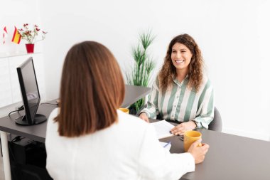 Two Caucasian women are discussing work matters in a sleek office environment. One woman holds a coffee cup while the other smiles, showcasing collaboration and professionalism. clipart