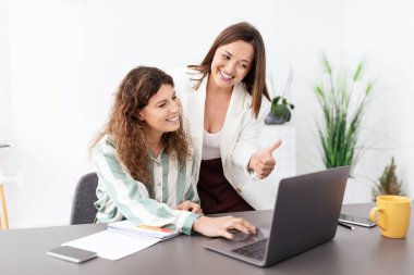 Çağdaş bir ofiste iki kadın bir tartışma içindeler. Biri dizüstü bilgisayarı işaret ederken diğeri gülümseyip etkileşime geçiyor, takım çalışması ve verimlilik gösteriyor..