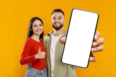 Genç bir çift, canlı sarı bir arkaplana karşı mutlu bir şekilde gülümsüyor. Erkek akıllı telefonu belirgin bir şekilde tutarken, kadın onların bağlantısını kutluyor..