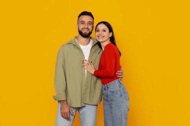 Genç bir çift, birbirlerine öylesine sarılırken geniş bir gülümseme sergiliyor. Parlak sarı arka plan, neşeli tavırlarını artırıyor ve neşeli bağlantılarını vurguluyor..