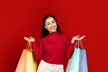 Kırmızı balıkçı yaka kazaklı genç bir beyaz kadın elinde renkli alışveriş torbaları tutarken geniş bir gülümsemeyle parlak kırmızı bir arka plan karşısında duruyor. Tüketim ve alışveriş sevincinin heyecanını somutlaştırıyor..