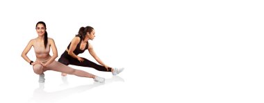 İki kadın dinamik esneme egzersizleri yapıyor, esneklik ve ısınmaya odaklanıyor. Rahat spor kıyafetleri giyiyorlar ve çeşitli esneme tekniklerini parlak bir alanda sergiliyorlar..