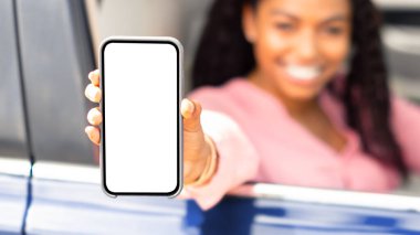 Gülümseyen siyahi kadının beyaz ekranlı cep telefonunu gösterip yeni lüks bir arabanın sürücü koltuğunda otururken, elinde aleti tutarken, cihaza odaklanırken, arka planda bulanıklık çekerken.