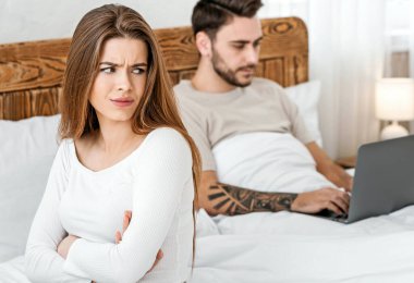 A lot of work concept. Man works on laptop, woman is offended by bed in bedroom. clipart